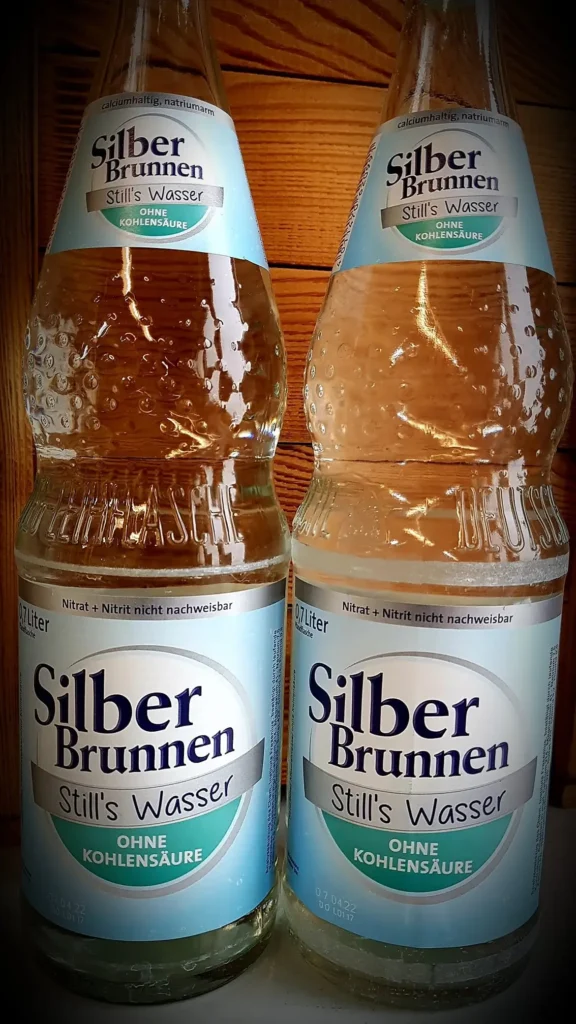 Zwei Flaschen mit blau-weißen Etiketten von stillem Wasser auf einem Tisch.